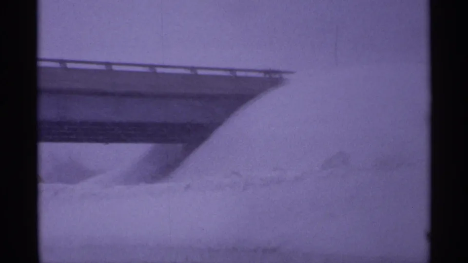 The frame centers on a large snowdrift beneath the overpass, with the guardrail and bridge deck partly visible through winter haze.