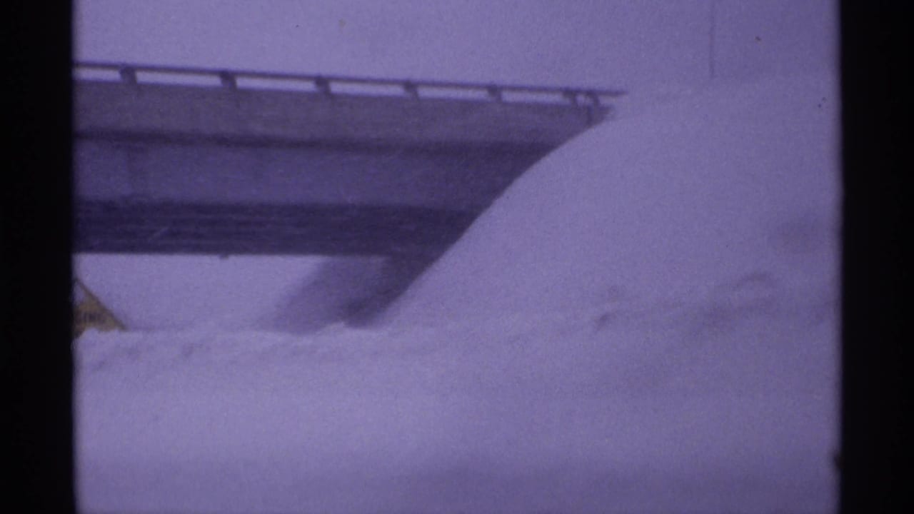 Deep snow piled beside a bridge or overpass with a yellow sign partly visible.