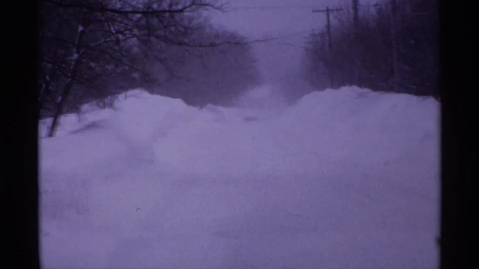 1971 Wisconsin home-movie frame showing a snowbound road with drifting snow, bare roadside trees, and utility poles.