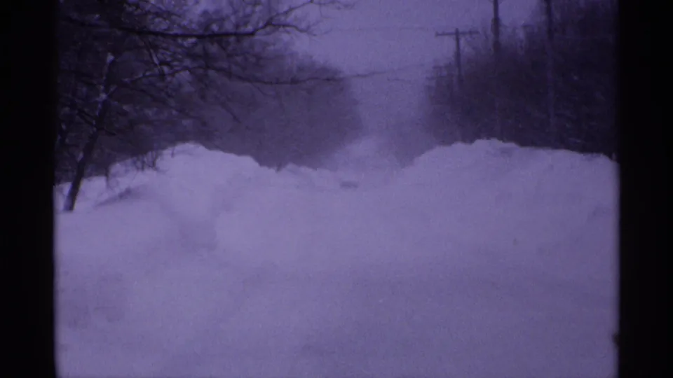 1971 Wisconsin home-movie frame showing deep snowdrifts across a yard with bare trees, utility poles, gray sky, and dark 8mm frame edges.