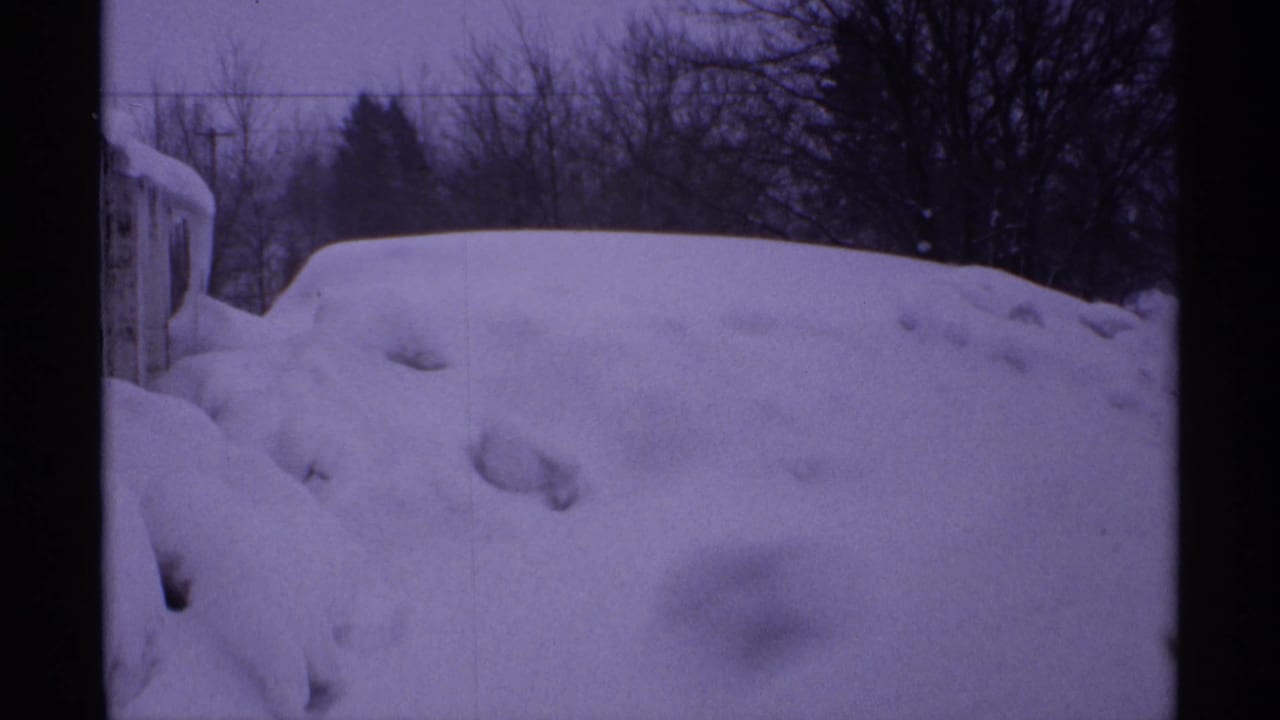 High snowbank beside a partially visible structure with bare trees in the background.