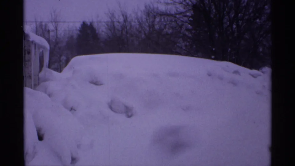 1971 Wisconsin winter frame with a tall snowbank, a partially visible building at left, bare trees, and utility lines in a purple-tinted 8mm scan.