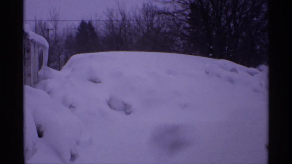 1971 Wisconsin home-movie view of deep snow piled across a residential yard, with a partial structure at left and winter trees beyond.