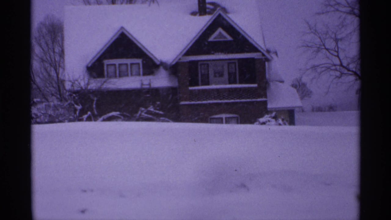 Snow-covered two-story house seen across a deep snowbank with bare trees behind it.