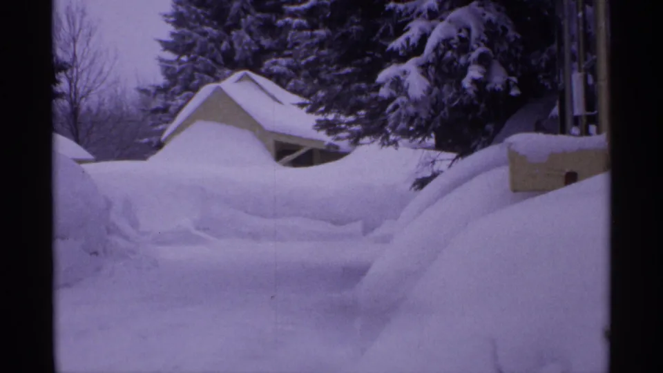 1971 Wisconsin home-movie frame of a small backyard shed surrounded by deep snow and heavy evergreen branches.