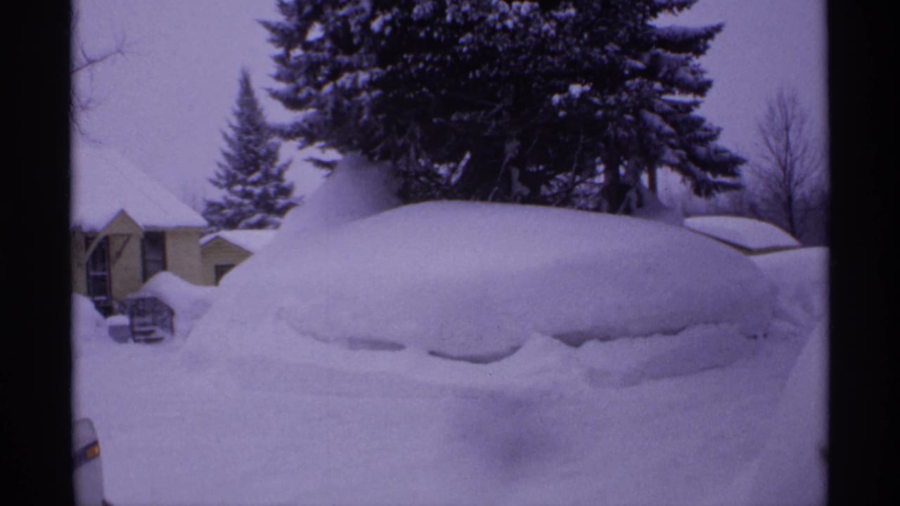 A car-shaped mound is buried under deep snow near houses and evergreens.