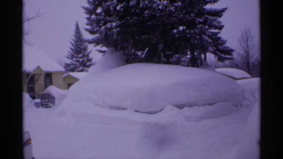 1971 Wisconsin home-movie frame with a heavily snow-covered car beside small houses and tall evergreens.