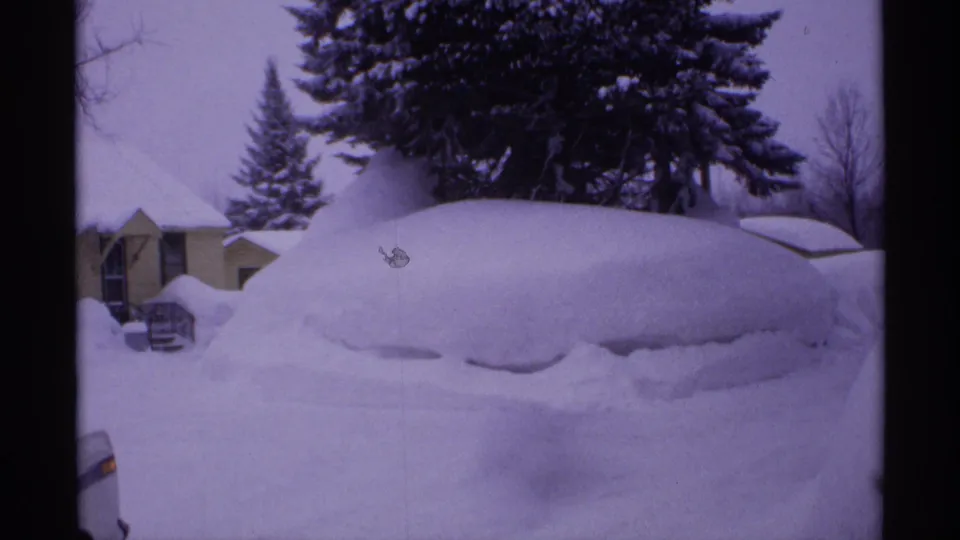 1971 Wisconsin winter frame showing a buried vehicle, nearby homes, and snow-heavy evergreens in cool 8mm color.