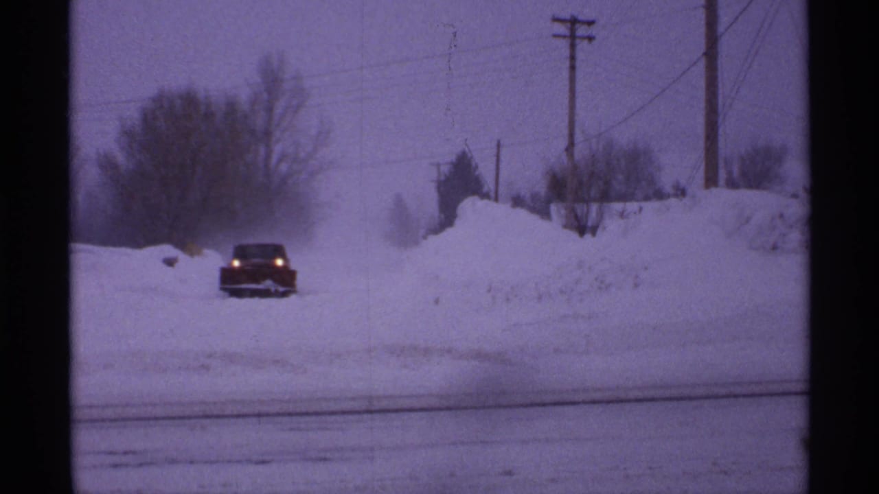 Red car with headlights approaches along a snowy road bordered by high snowbanks.