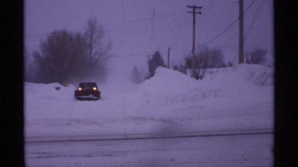 1971 Wisconsin 8mm frame showing a red car approaching through snow on a road lined with high banks, utility poles, and bare trees.