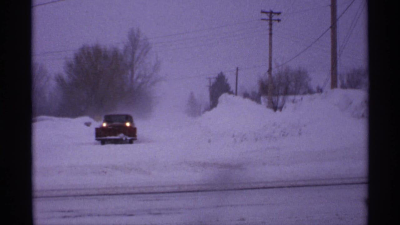 1971 Wisconsin frame of a red car driving through drifting snow, with tall roadside snowbanks and utility poles visible in the background.