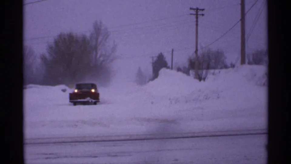 1971 Wisconsin frame of a red car driving through drifting snow, with tall roadside snowbanks and utility poles visible in the background.