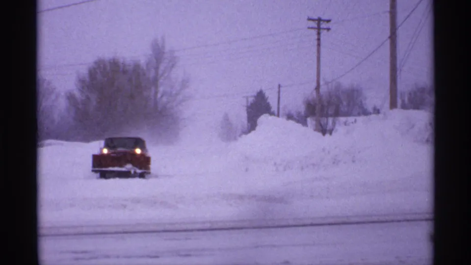 1971 Wisconsin 8mm frame showing the red car closer on the snow-covered road, with bare trees and utility poles beyond the drifts.