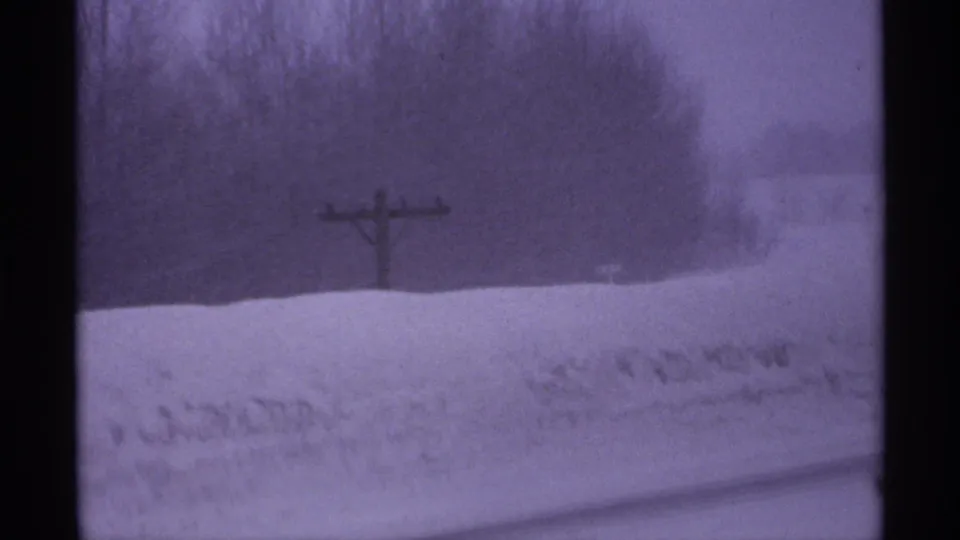 1971 Wisconsin 8mm frame showing the snowy roadside after the car passes, with a utility pole, snowbank, bare trees, and blowing snow.
