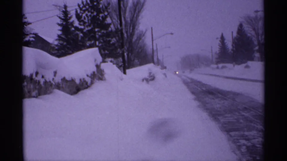 1971 Wisconsin 8mm frame showing a snow-lined residential street, evergreens, a stone wall, utility lines, and distant car headlights.