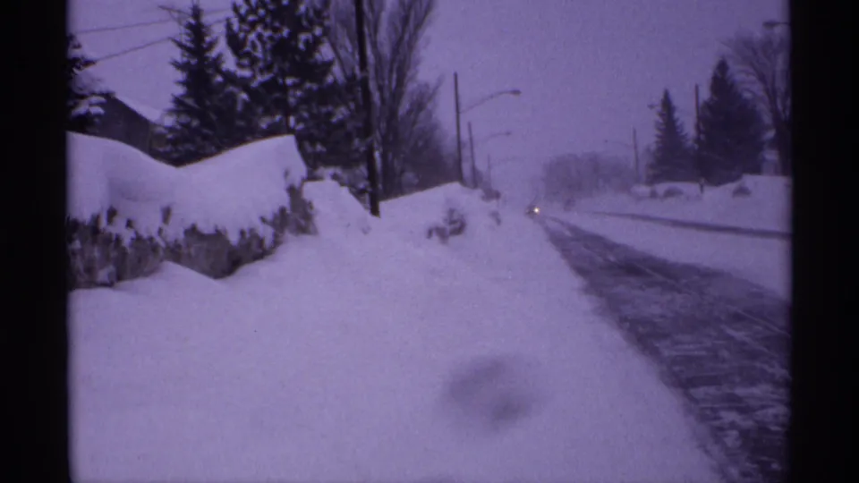 1971 Wisconsin home-movie frame of a snowy residential road with bare trees, evergreens, utility poles, and a small distant headlight.