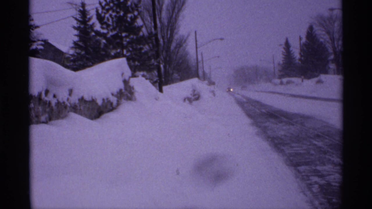 Snow-covered roadside with high banks, trees, utility poles, and distant vehicle headlights.