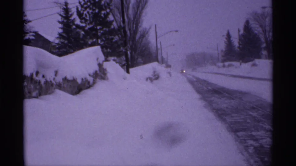 1971 Wisconsin 8mm frame showing a snow-lined road, tall plowed banks, bare trees, evergreens, utility poles, and distant headlights.