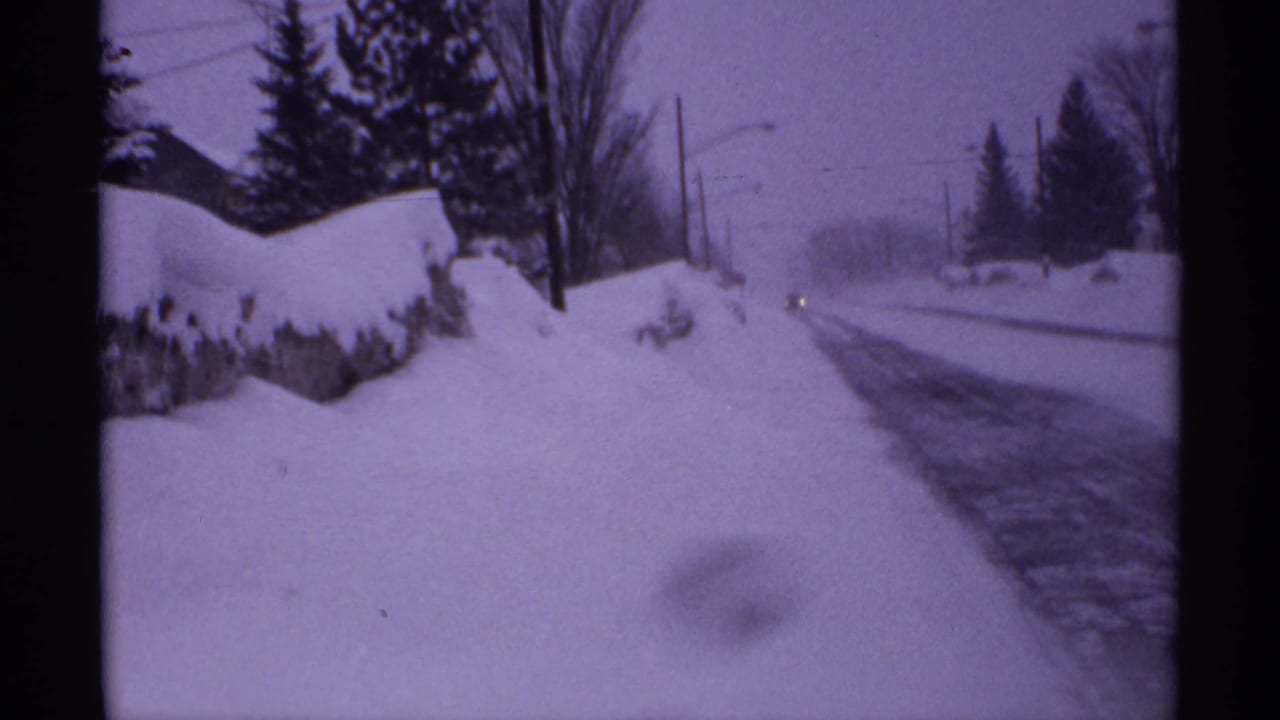 Snowy roadside with plowed banks, trees, utility poles, and a faint vehicle light ahead.