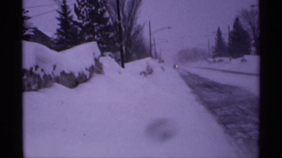 1971 Wisconsin winter frame with a snowbank beside a partially plowed road, evergreens, bare trees, utility poles, and distant headlights.