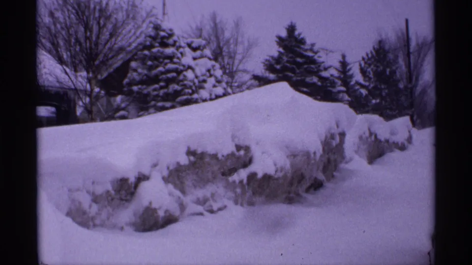 1971 Wisconsin 8mm frame showing heavy snow built up over a low stone or retaining wall, with evergreens and leafless trees behind.