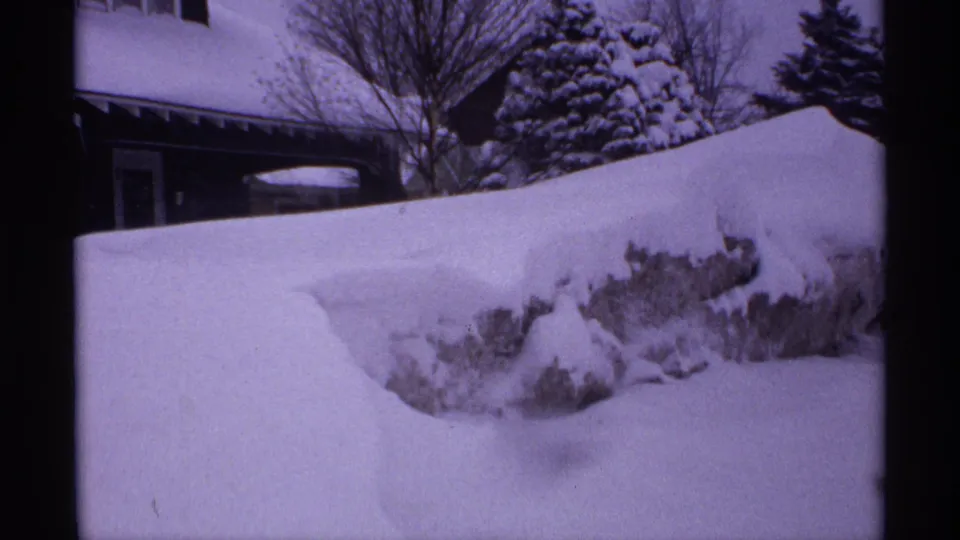 Wisconsin 1971 home-movie frame showing a deep snowbank, house exterior, bare trees, and snow-covered evergreens.