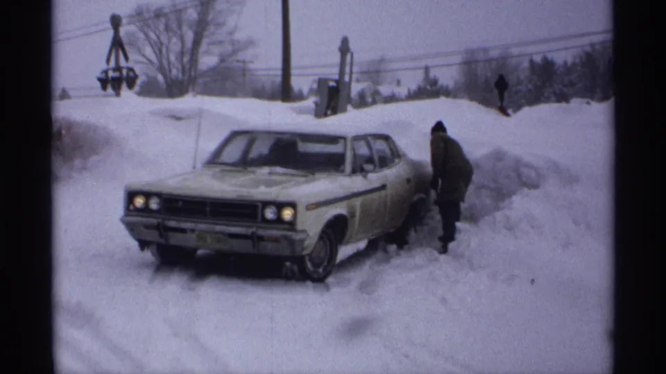A man in winter clothing shovels around a white sedan stuck in deep snow in a Wisconsin neighborhood, 1971.
