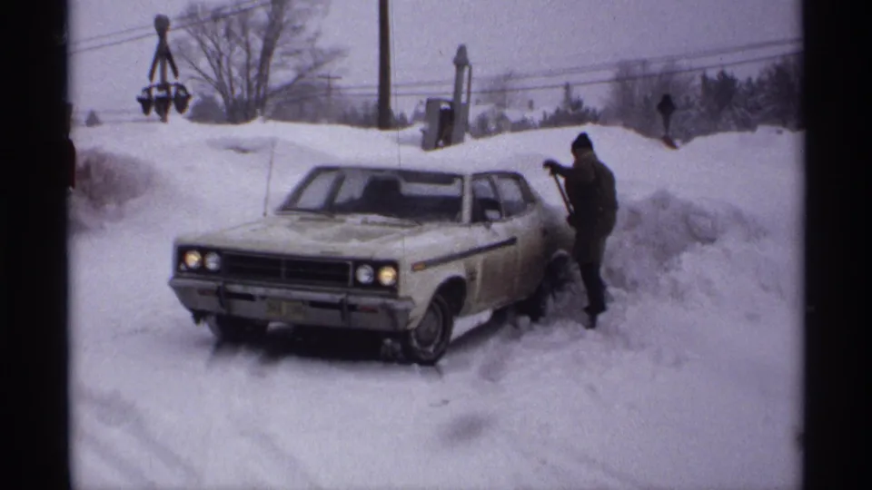 Wisconsin 1971 footage shows a man shoveling near a white sedan, with snowbanks, trees, and utility lines behind him.
