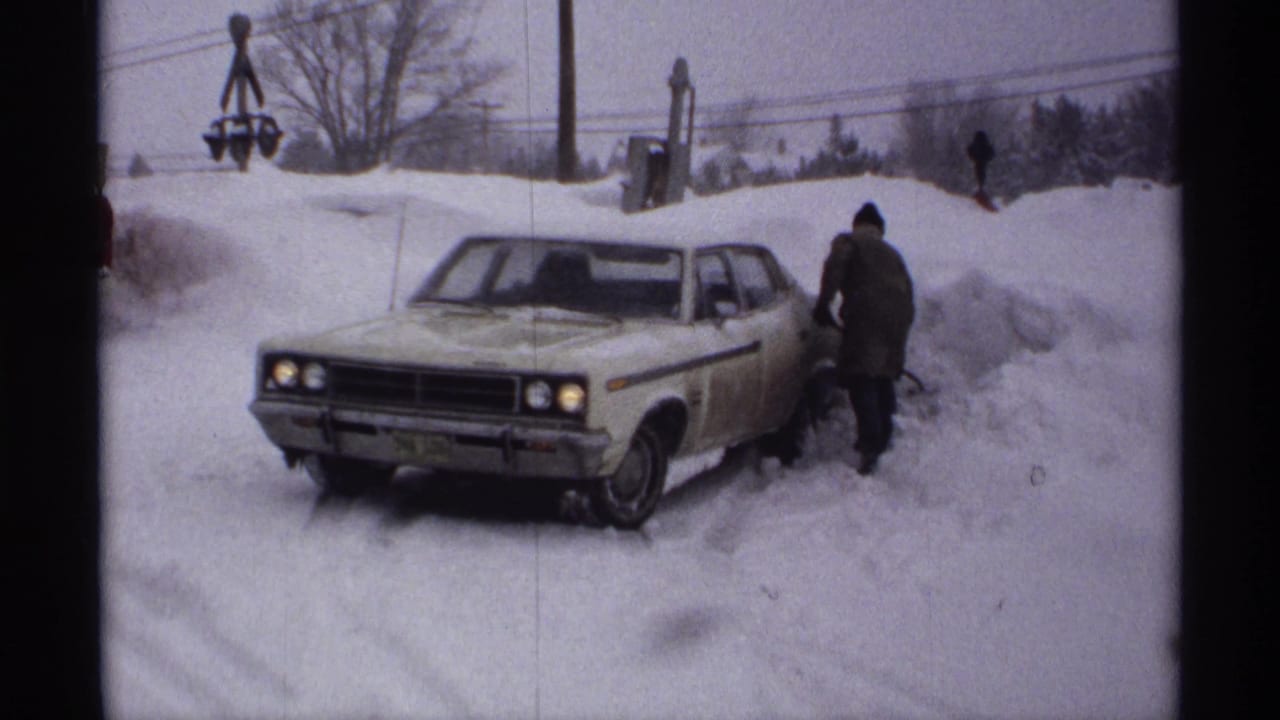 Light-colored sedan stuck in deep snow with a bundled person beside it.
