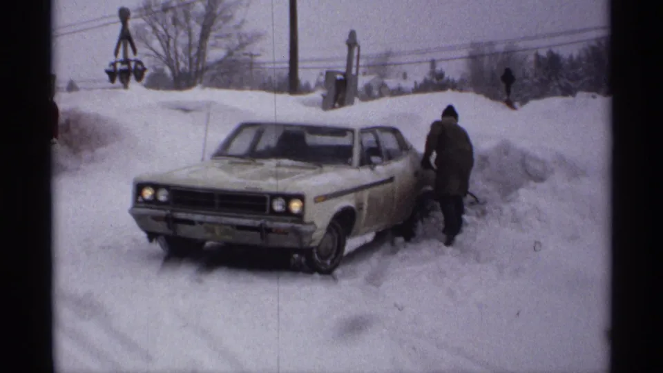 1971 Wisconsin 8mm frame showing a light-colored sedan in heavy snow as a bundled adult stands near the passenger side.