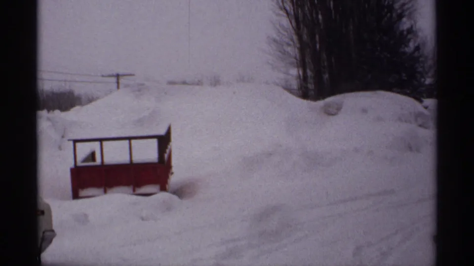 A 1971 Wisconsin winter frame shows deep snow piled around a small red roadside structure, with utility lines and trees beyond.