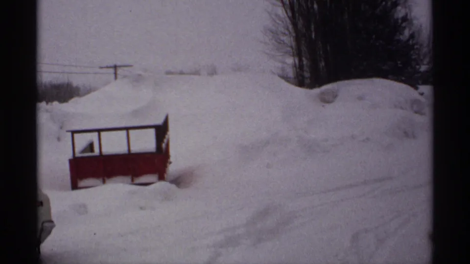 The rural Wisconsin roadside is buried under heavy snow, with a red structure partly framed at lower left and trees in the background.