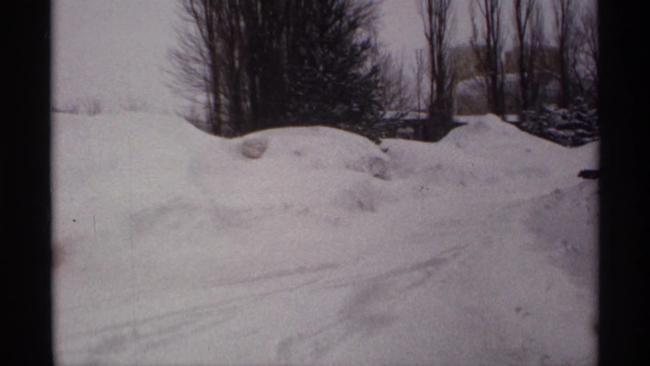 1971 Wisconsin 8mm frame of snowdrifts, faint tire tracks, bare trees, and overhead power lines under a gray winter sky.