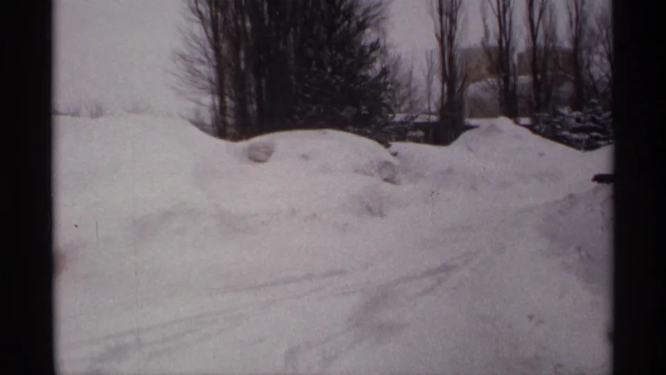 1971 Wisconsin 8mm frame of snowdrifts, faint tire tracks, bare trees, and overhead power lines under a gray winter sky.