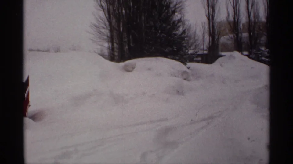 1971 Wisconsin 8mm frame of snowdrifts, faint vehicle tracks, bare trees, and a sliver of red trailer at the edge.