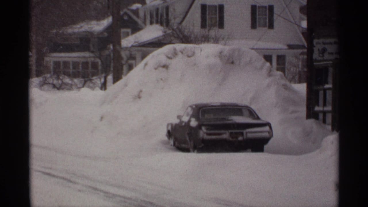 1971 Wisconsin home-movie frame showing a dark sedan nearly surrounded by deep snow on a residential street.