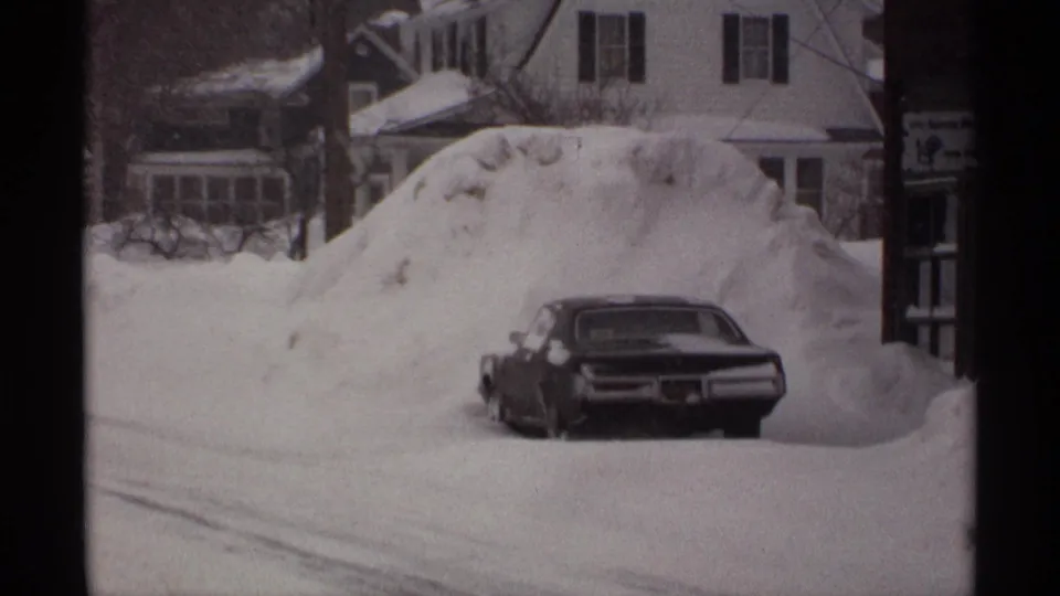 1971 Wisconsin home-movie frame showing a dark sedan nearly surrounded by deep snow on a residential street.