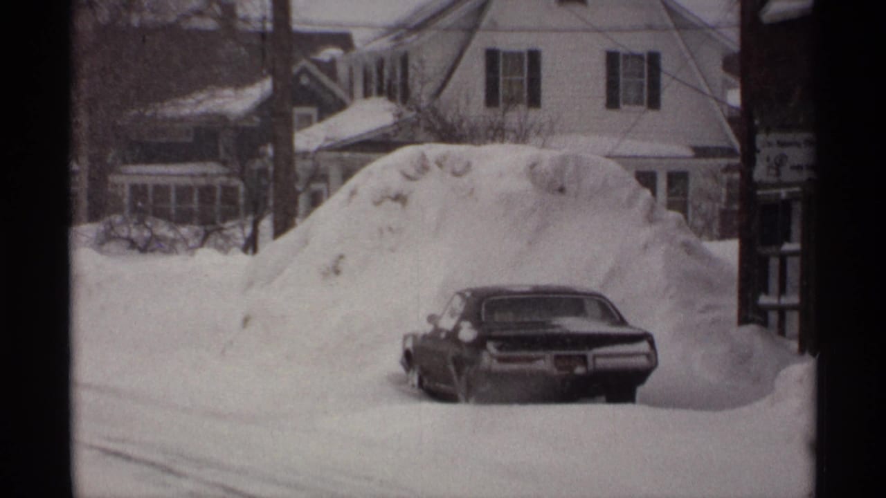 Parked car beside a large snowdrift with houses and utility wires behind it.