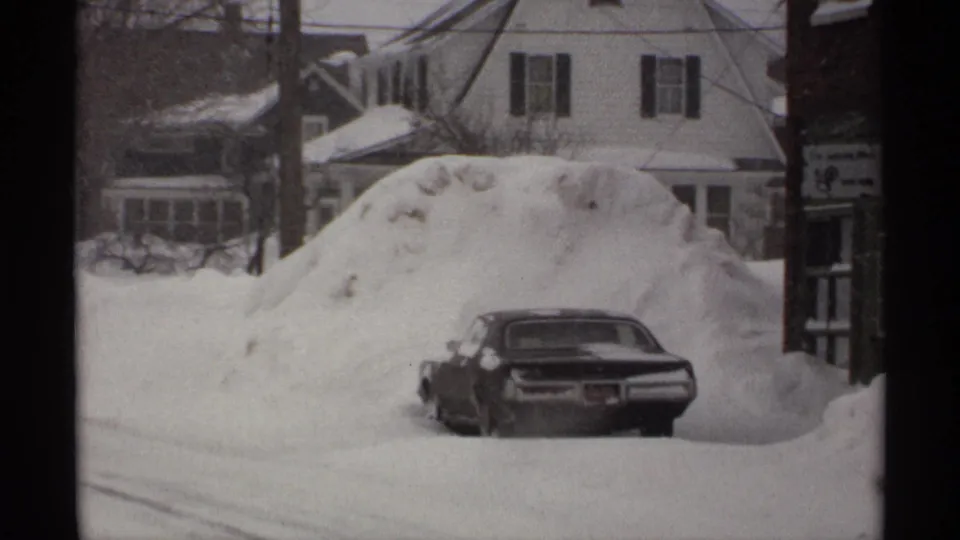 A 1971 Wisconsin winter frame shows the rear of a dark sedan tucked into deep snow near homes with icicles.