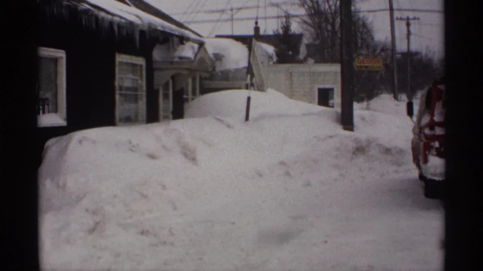 The camera shifts along the Wisconsin street to show high snowbanks, icicles on a dark building, utility poles, and a partial vehicle.