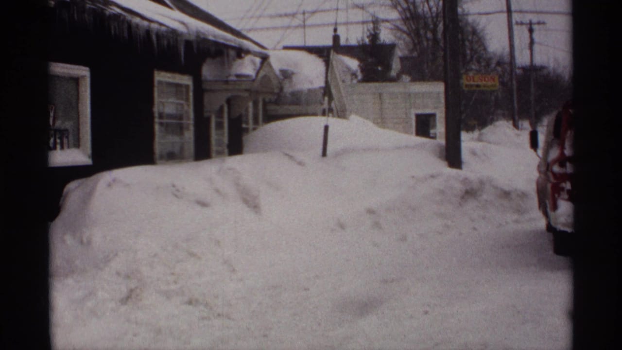 1971 Wisconsin home-movie frame showing deep snowbanks, icicles on eaves, older houses, utility wires, and a roadside sign.