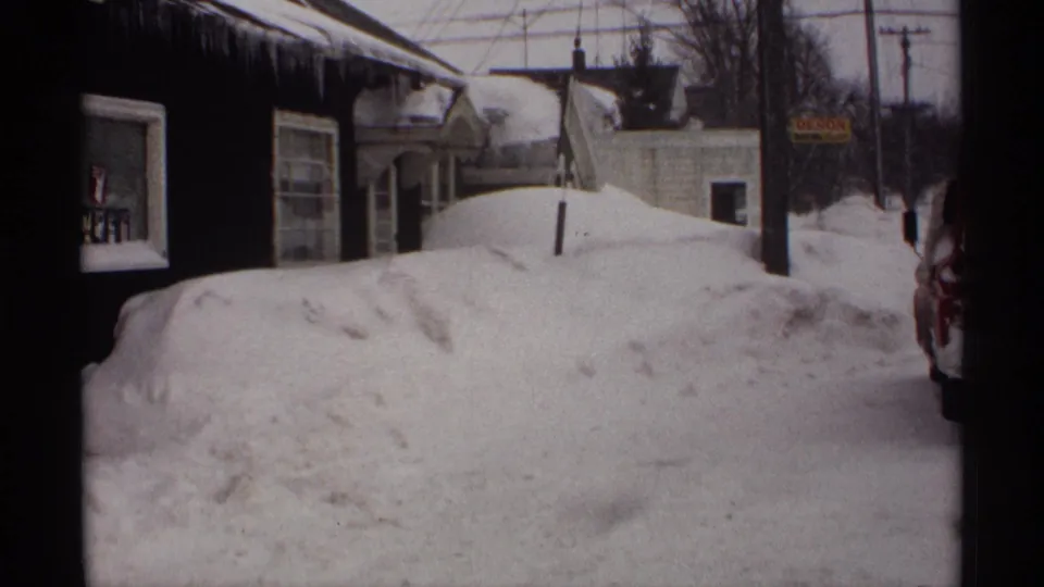 1971 Wisconsin frame with snow piled high outside residential buildings, icicles under the eaves, and a small sign near the road.