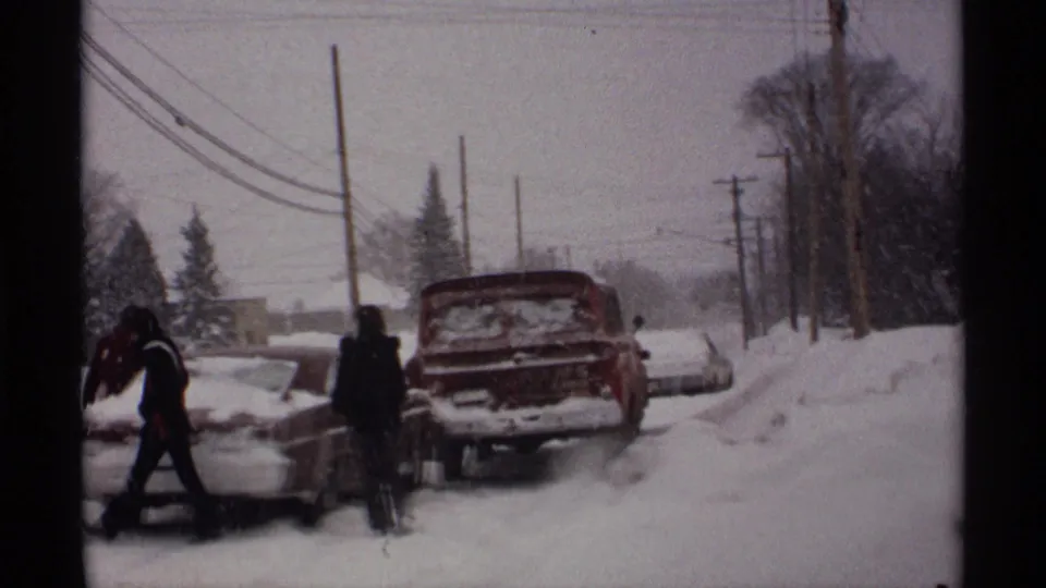 Wisconsin winter home-movie frame from 1971 showing people clearing snow from parked cars near a red truck and utility poles.