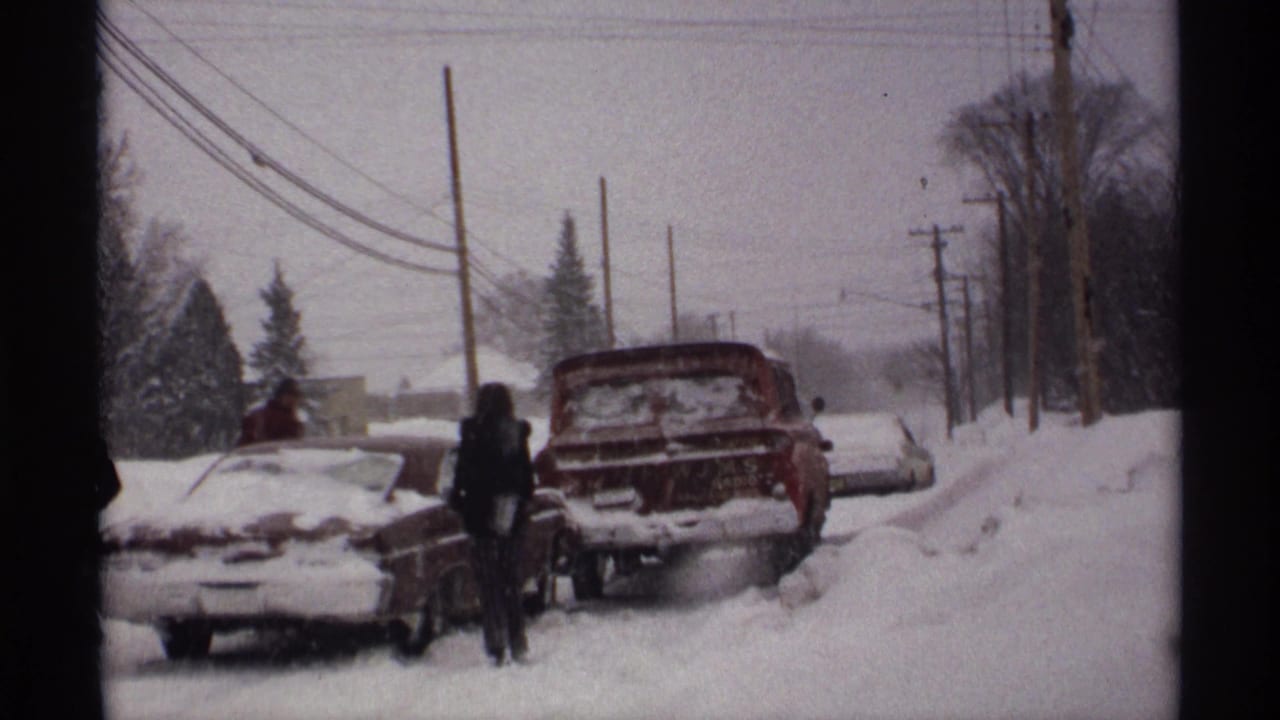 Snow-covered suburban street with two cars, bundled people, utility poles, and deep roadside snow.