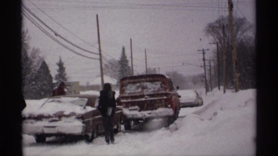 1971 Wisconsin home-movie frame showing a snowbound suburban street with buried cars, bundled people, utility poles, and winter trees.