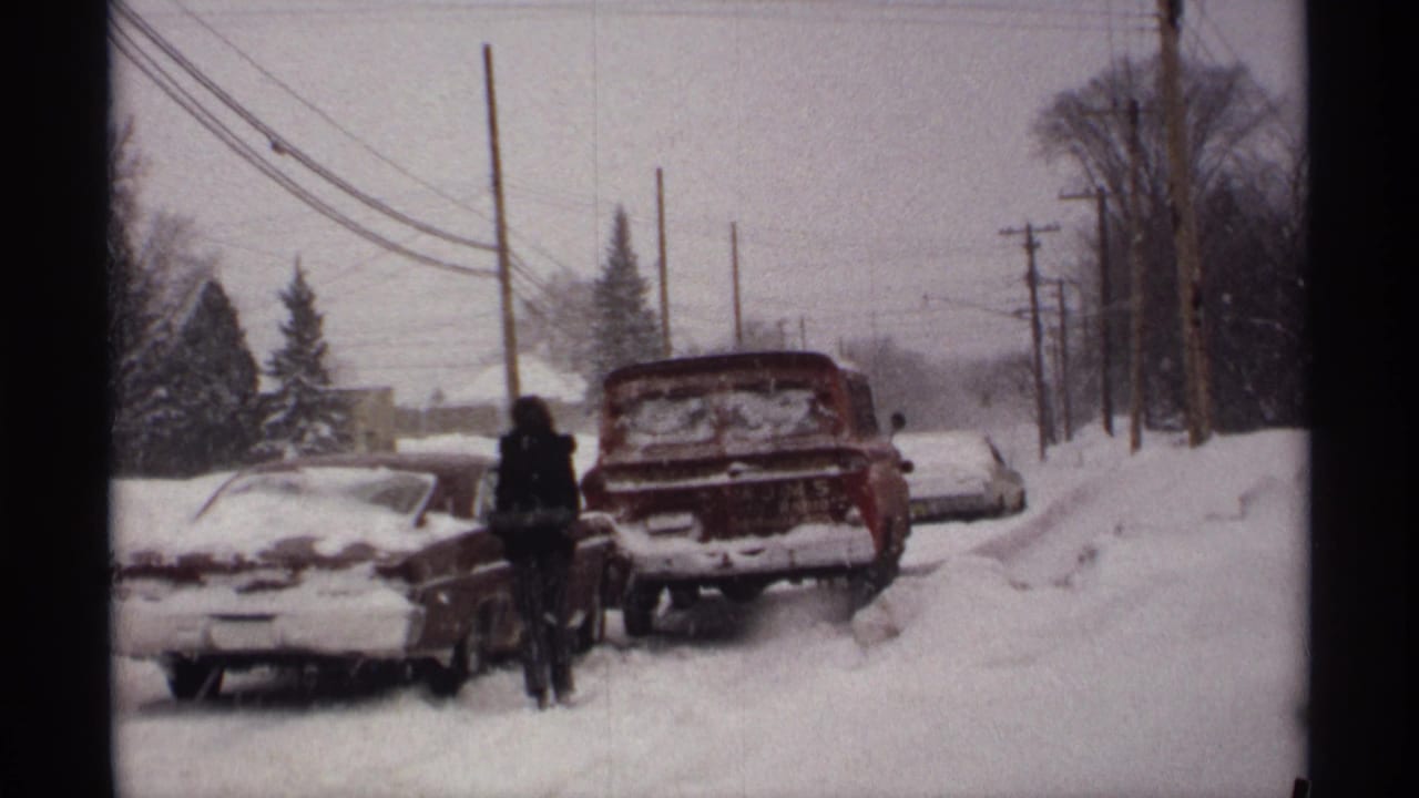 Two snow-covered cars sit along a snowy road while bundled figures stand nearby.