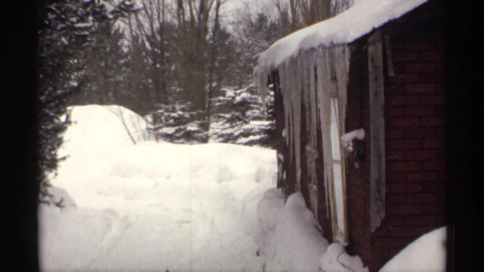 1971 Wisconsin home-movie view of a snow-covered yard beside a red building, with evergreens, bare trees, and deep drifts.