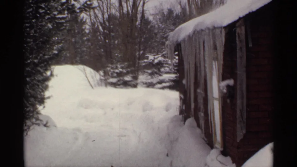 1971 Wisconsin frame showing the side of a red building, heavy snowdrifts, evergreens, and leafless trees in a residential winter setting.