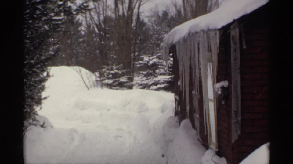 1971 Wisconsin 8mm frame showing deep snow beside a brown building, with long icicles, evergreens, and bare trees in the background.