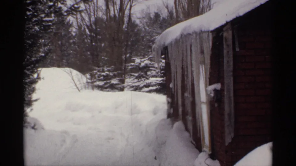 1971 Wisconsin winter frame with icicles hanging from a brown building, deep snow in the yard, and trees beyond the structure.